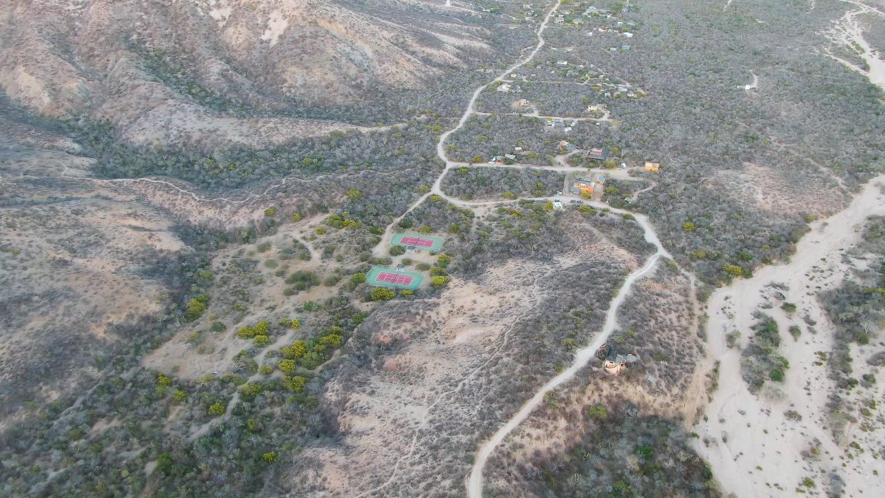 Aerial view in Cabo Pulmo with the dirt road network more centered, small structures near the upper slope, bright rectangular courts below, and no visible people.