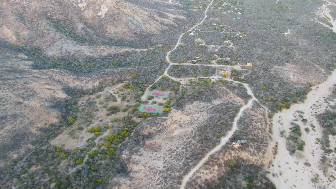 High aerial frame over Cabo Pulmo with the dry wash running through the right side, scrub-covered slopes, small hillside buildings, and no visible people.
