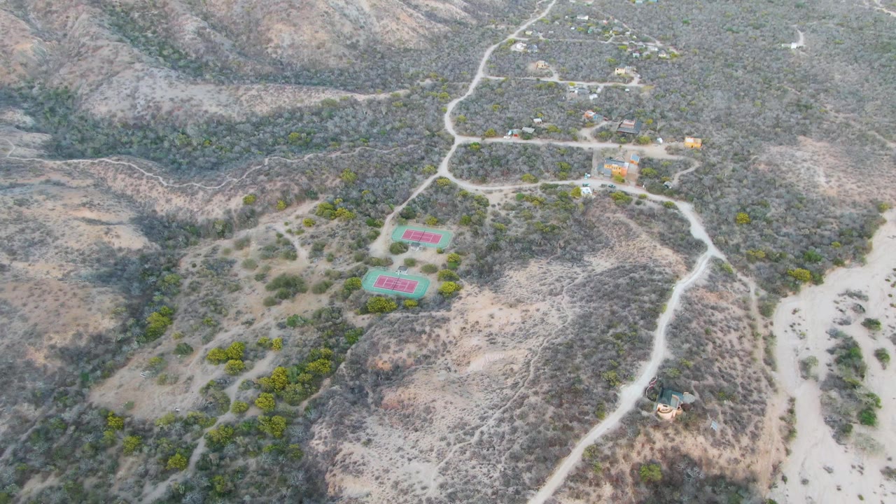 Opening aerial frame over two red and green tennis courts in Cabo Pulmo, Baja California Sur, surrounded by dry hills, pale dirt roads, scrub vegetation, and no visible people.