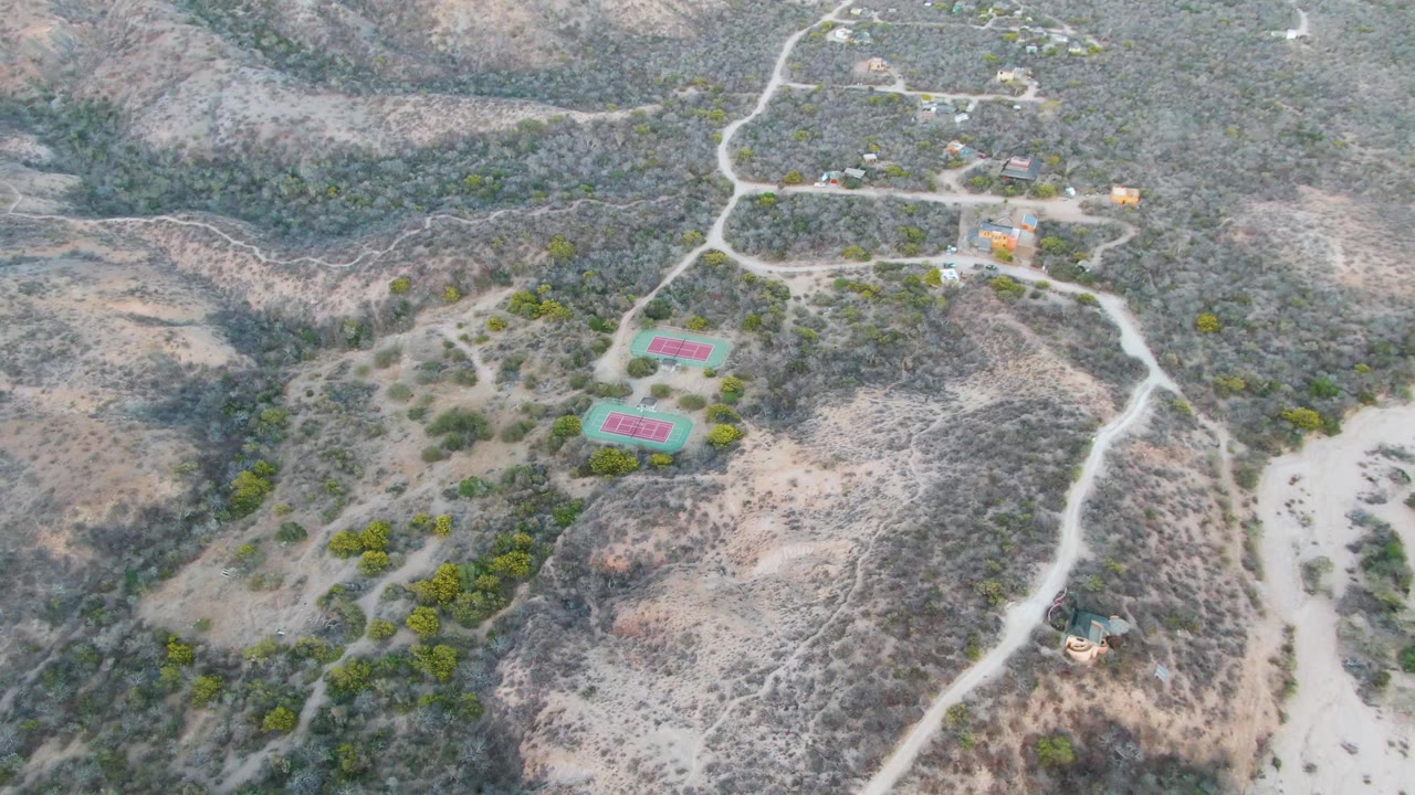Aerial view one second into the clip shows the Cabo Pulmo tennis courts slightly more centered between winding dirt roads, dry slopes, sparse shrubs, and no visible people.