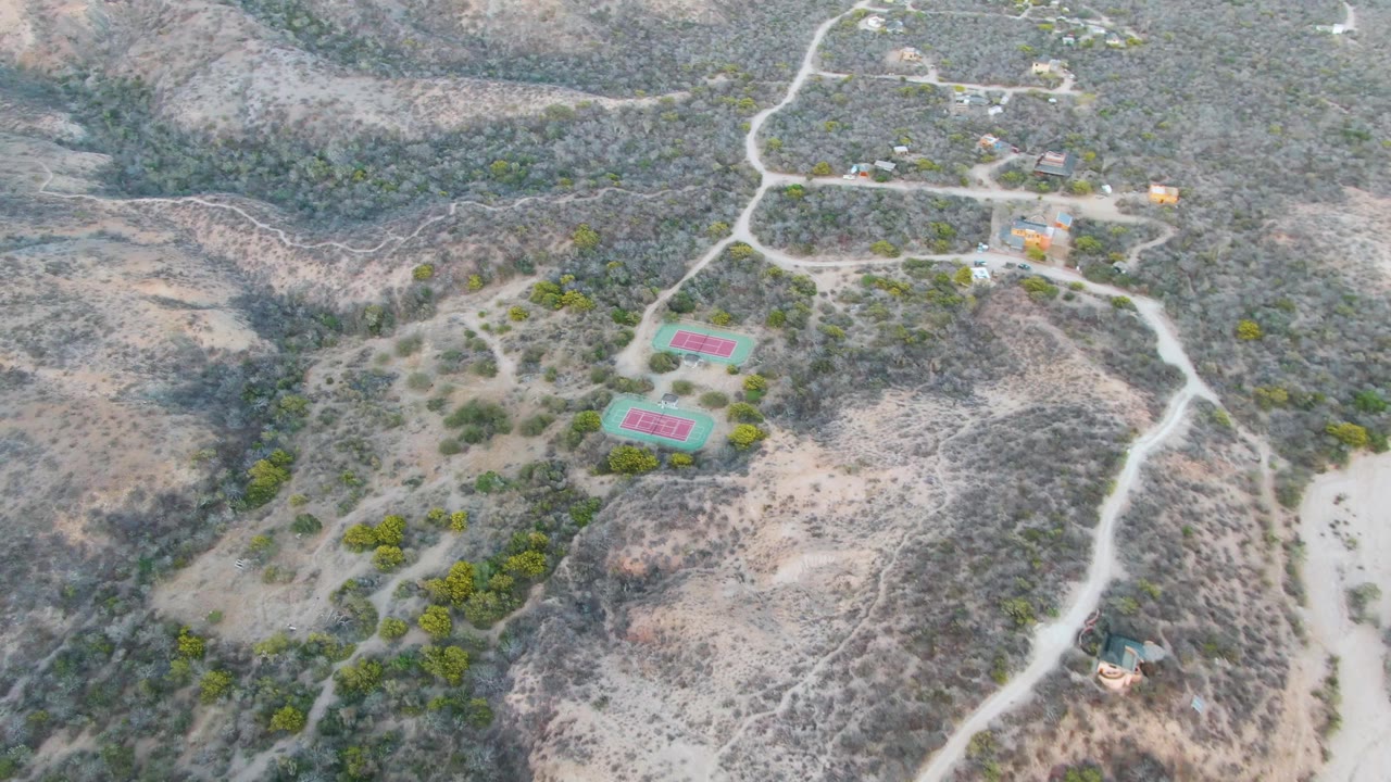 High aerial frame over Cabo Pulmo shows the two colored tennis courts below a cluster of small structures, with dusty tracks, patchy green shrubs, and no visible people.