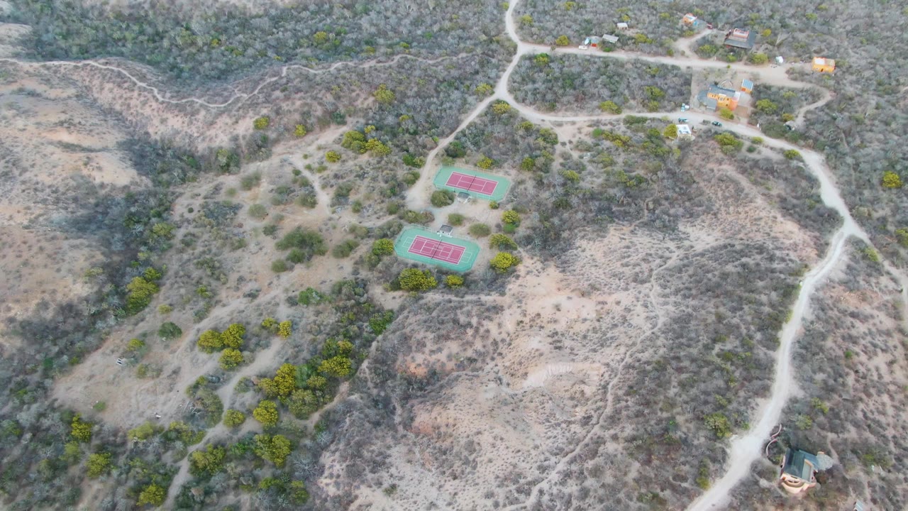 High aerial view over two red and green tennis courts on a dry Cabo Pulmo hillside, with winding dirt roads, scrub vegetation, scattered trees, small buildings, and no visible people.