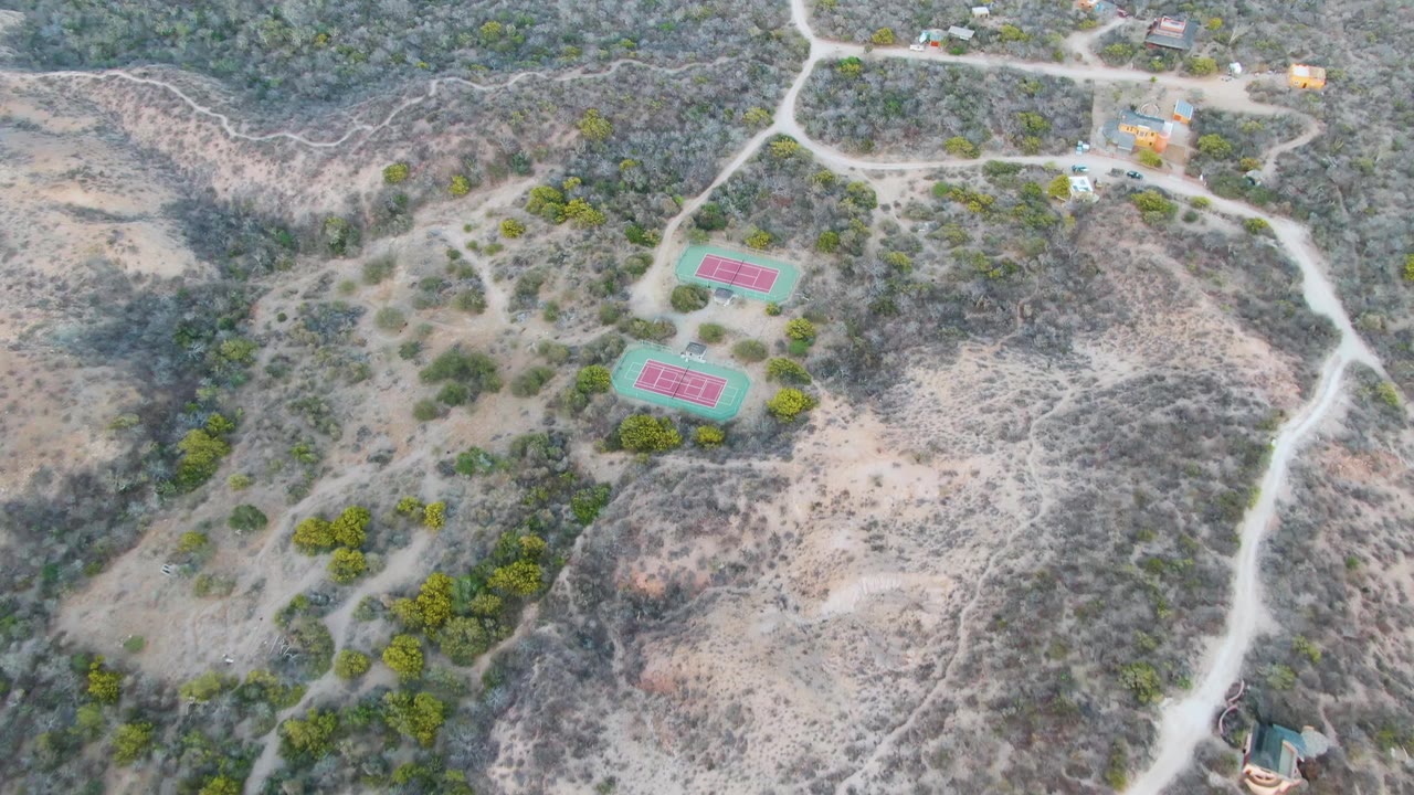 Drone view in Cabo Pulmo showing the pair of tennis courts slightly left of center, bordered by pale paths, dry gullies, scrub, trees, and distant small structures, with no visible people.