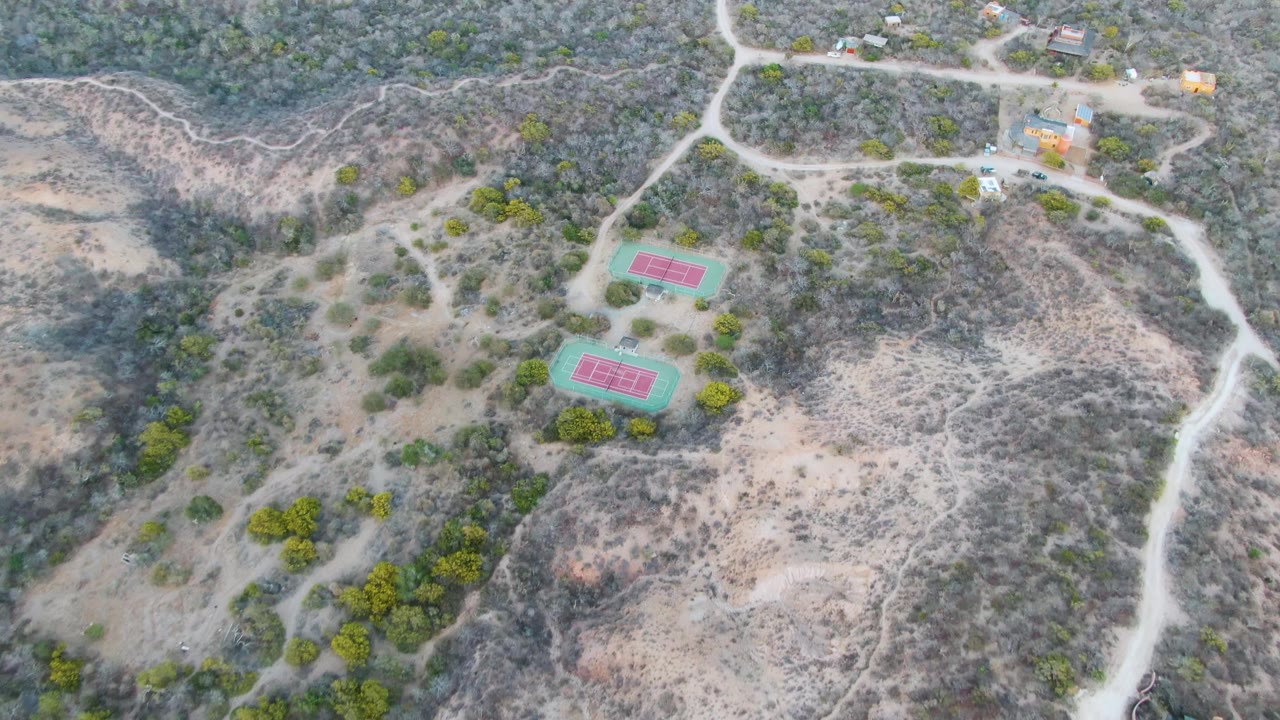 Aerial frame over Cabo Pulmo dry terrain with two red courts inside green surrounds, a main pale road curving along the right side, scrub-covered slopes, and no visible people.