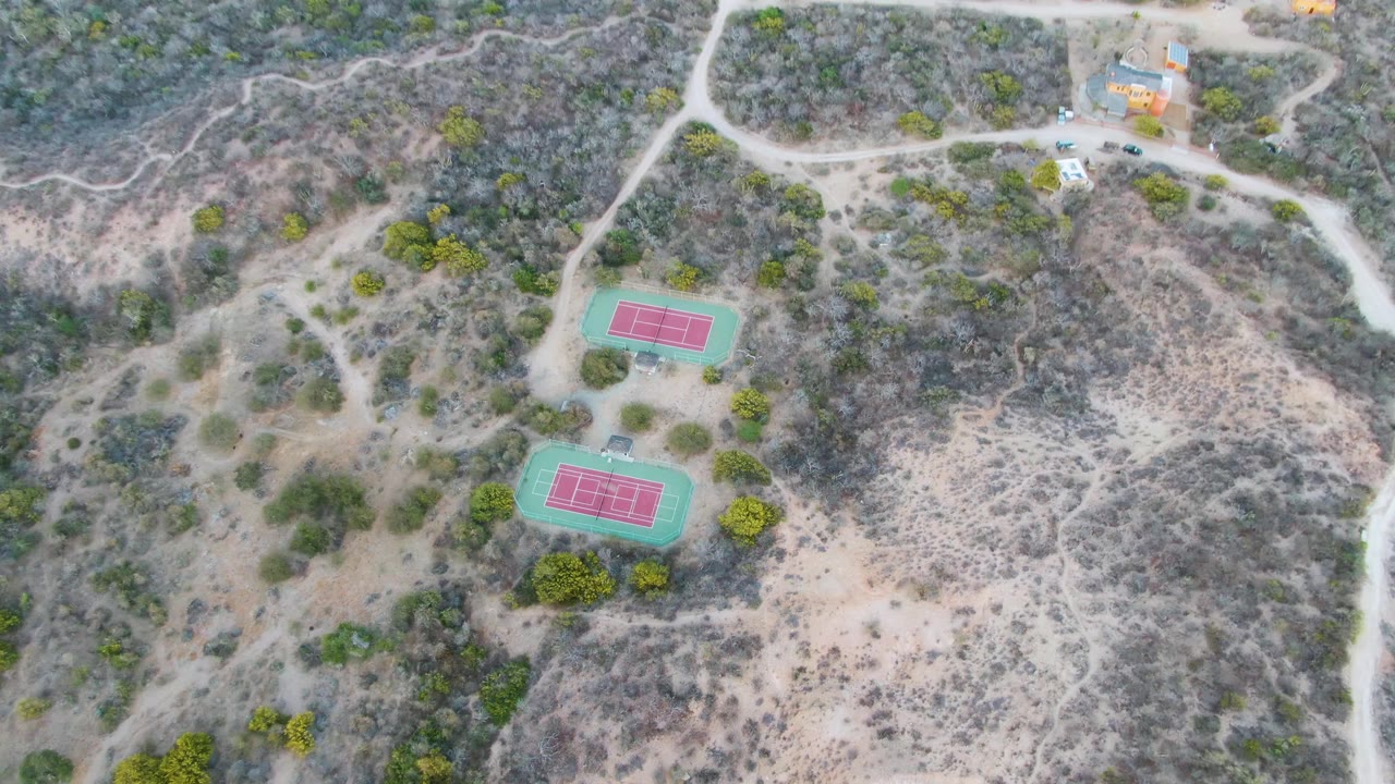 Aerial frame over Cabo Pulmo, Baja California Sur with the two tennis courts lower in view, dusty roads curving above them, scattered vehicles, and no visible people.
