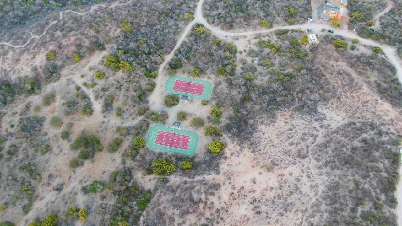 High drone view in Cabo Pulmo, Baja California Sur showing the paired courts near the lower left, scrubby hills to the right, pale tracks, vehicles, and no visible people.