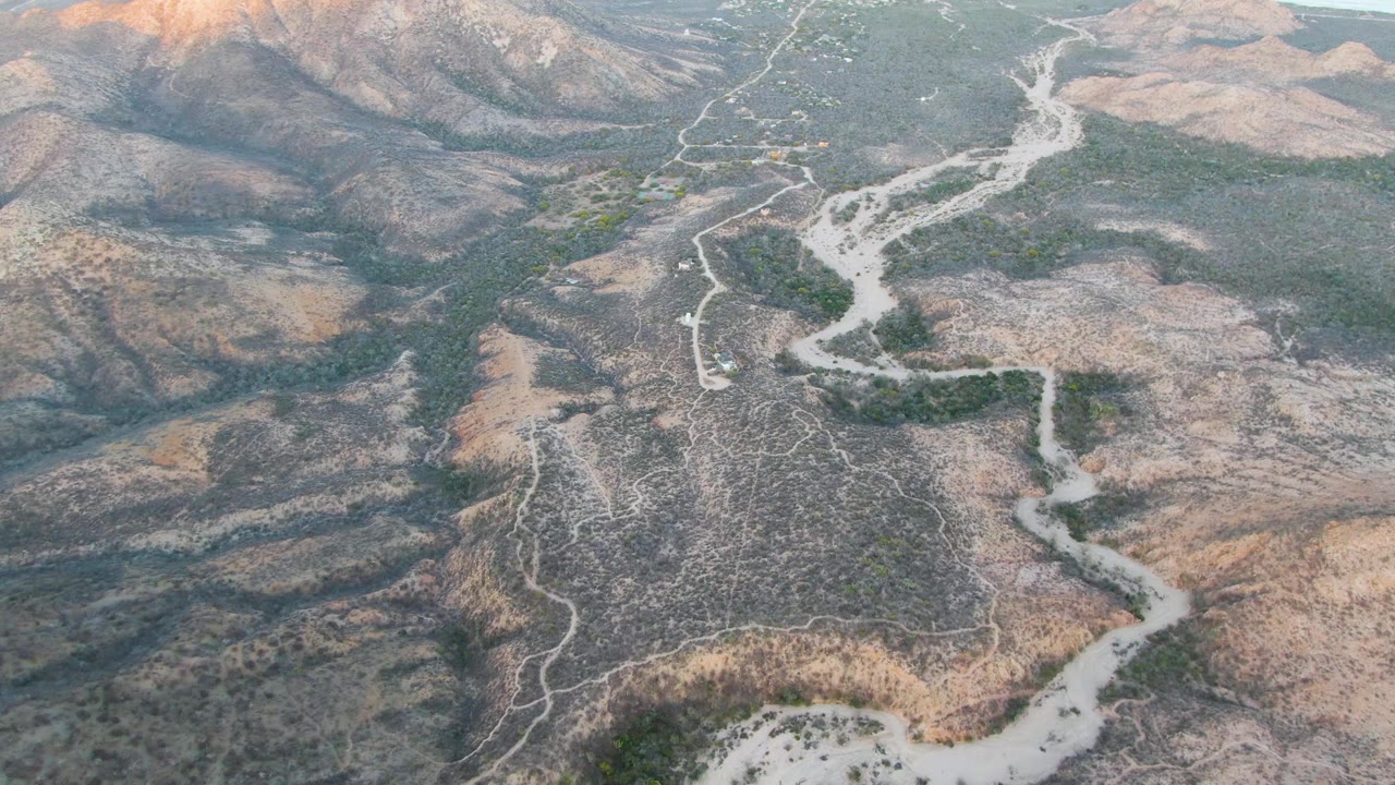 High aerial view of a winding dirt road through dry hills with a small vehicle visible.