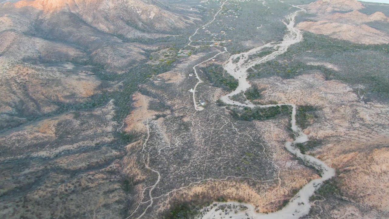Drone-height view near Cabo Pulmo with the dirt road bending through arid brown terrain, green patches along the wash, distant ridges, and no visible people.