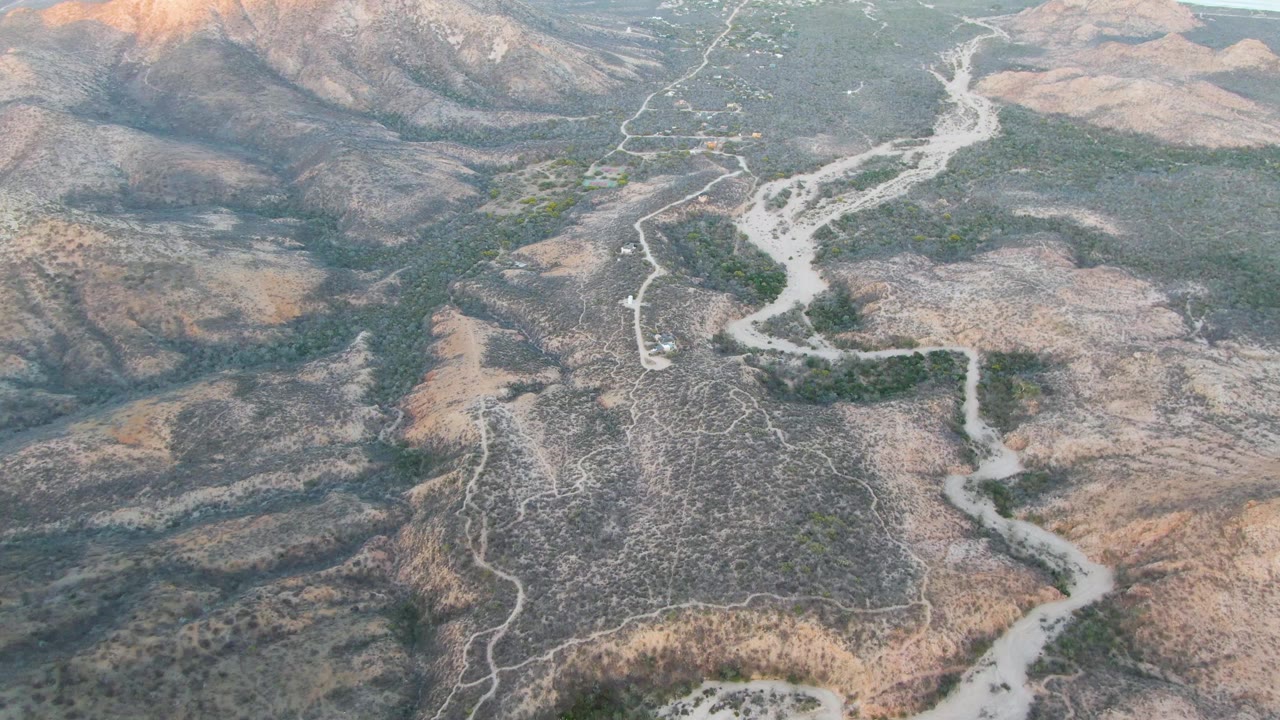 Aerial frame near Cabo Pulmo showing the winding dirt road running through dry Baja California Sur hills, with low green vegetation following the wash and no visible people.