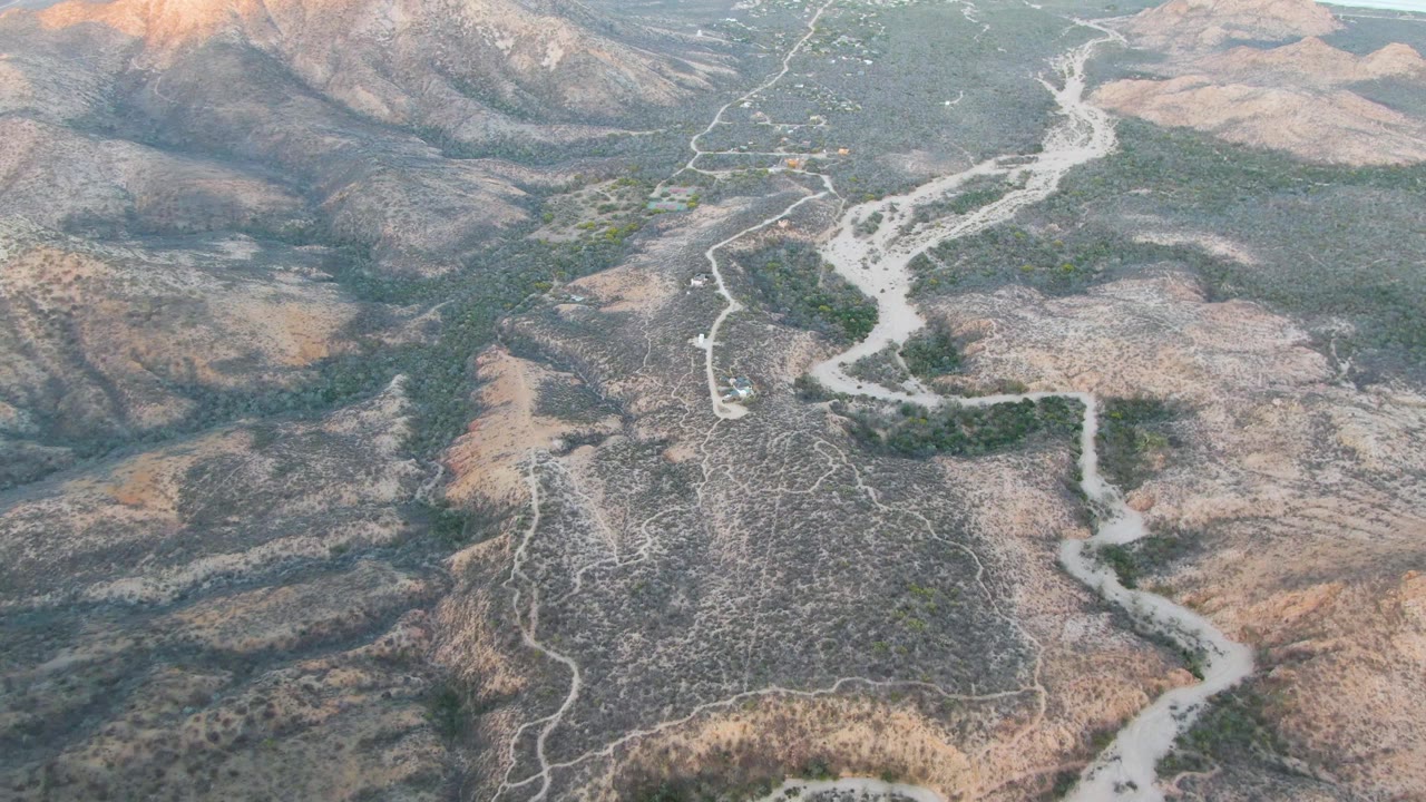 High drone perspective near Cabo Pulmo with the pale road and dry wash cutting through brown ridges, scattered shrubs, a small vehicle on the route, and no visible people.
