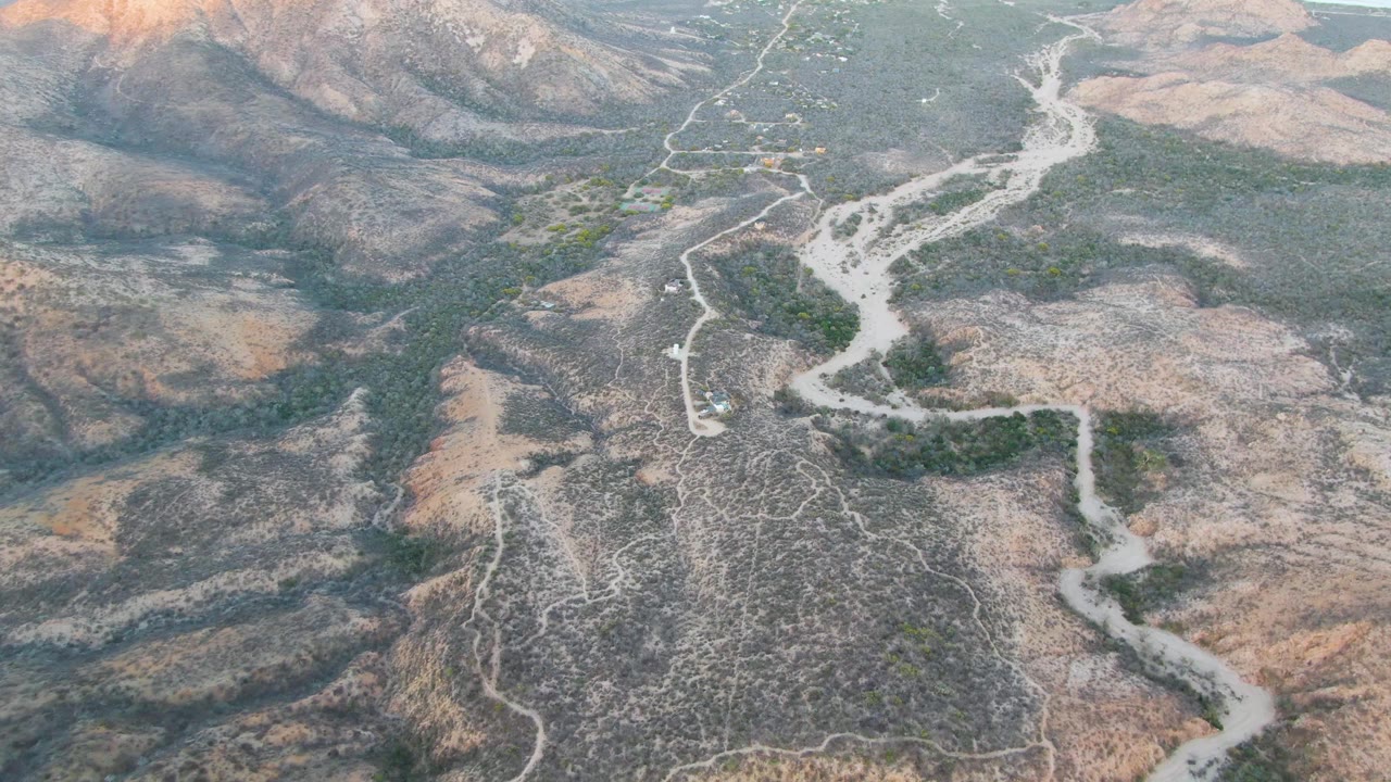 Aerial frame over Cabo Pulmo arid ridges, with the pale wash running through the center, green patches along the drainage, dirt paths, and no visible people.