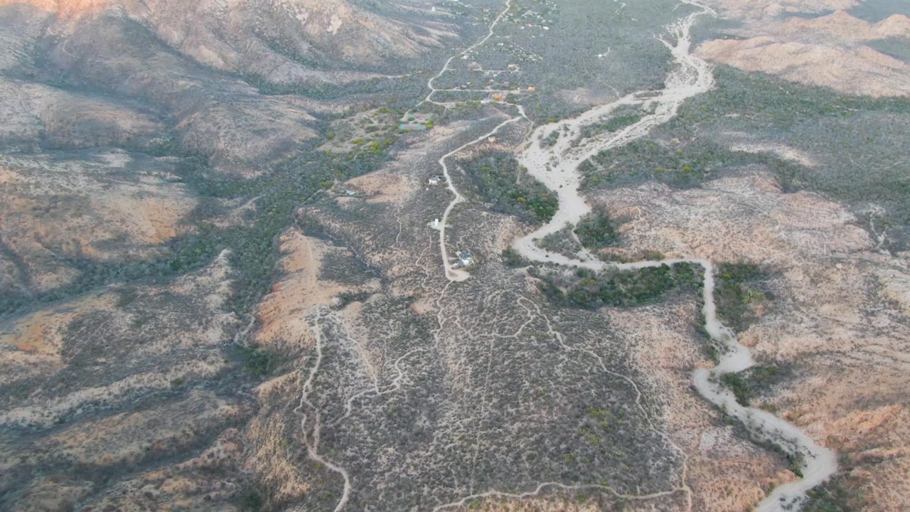Drone frame over Cabo Pulmo dry valley terrain, showing branching dirt tracks, a pale wash through the midground, gray-brown slopes, sparse vegetation, and no visible people.