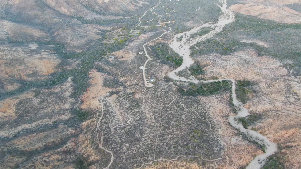 Aerial frame over Cabo Pulmo with the pale wash more prominent through the center, dry hills on both sides, isolated green vegetation, dirt paths, and no visible people.