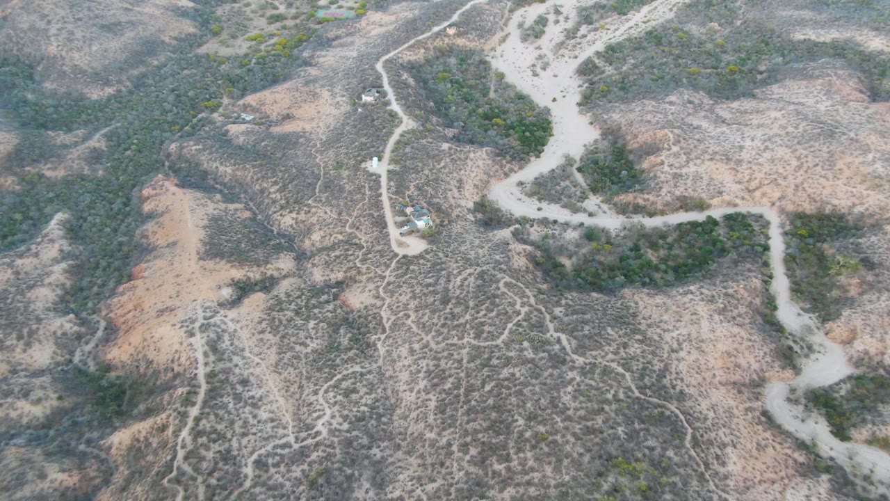 High drone frame from Cabo Pulmo showing the road snaking past sparse vegetation and small light-colored structures in an arid hillside setting, with no visible people.