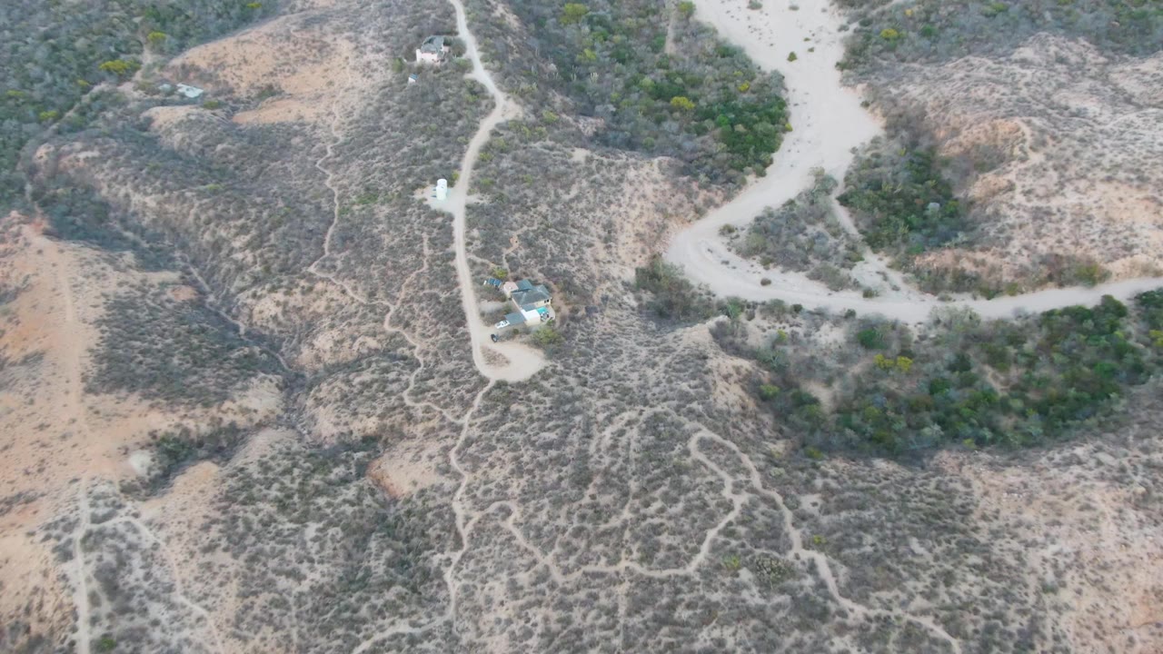 Overhead drone frame in Cabo Pulmo, Baja California Sur, showing a small vehicle near a pale dirt road, dry scrub hills, scattered structures, and no visible people.