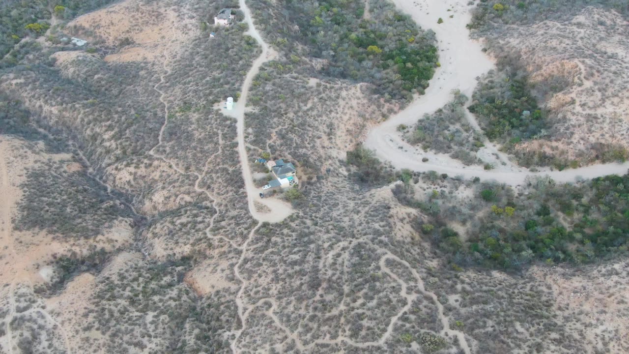 Overhead view in Cabo Pulmo showing the pale road curving past dry vegetation, small distant structures, earth-toned slopes, and no visible people.