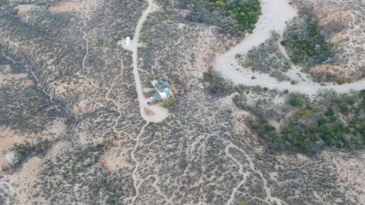 Aerial view over Cabo Pulmo arid scrubland with pale dirt roads, a small structure cluster left of center, green brush patches, and no visible people.