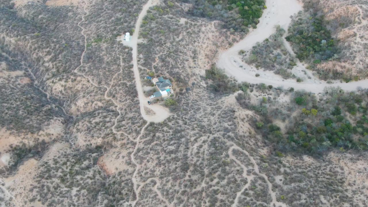 Top-down drone frame in Cabo Pulmo showing dry rocky terrain, branching pale roads, a compact group of small buildings near the left edge, and no visible people.