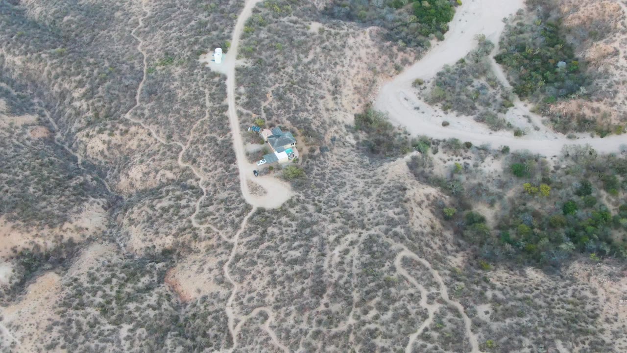 Drone view over Cabo Pulmo dry hills where a pale dirt road curves past small light-roofed structures, surrounded by scrub and darker green vegetation, with no visible people.
