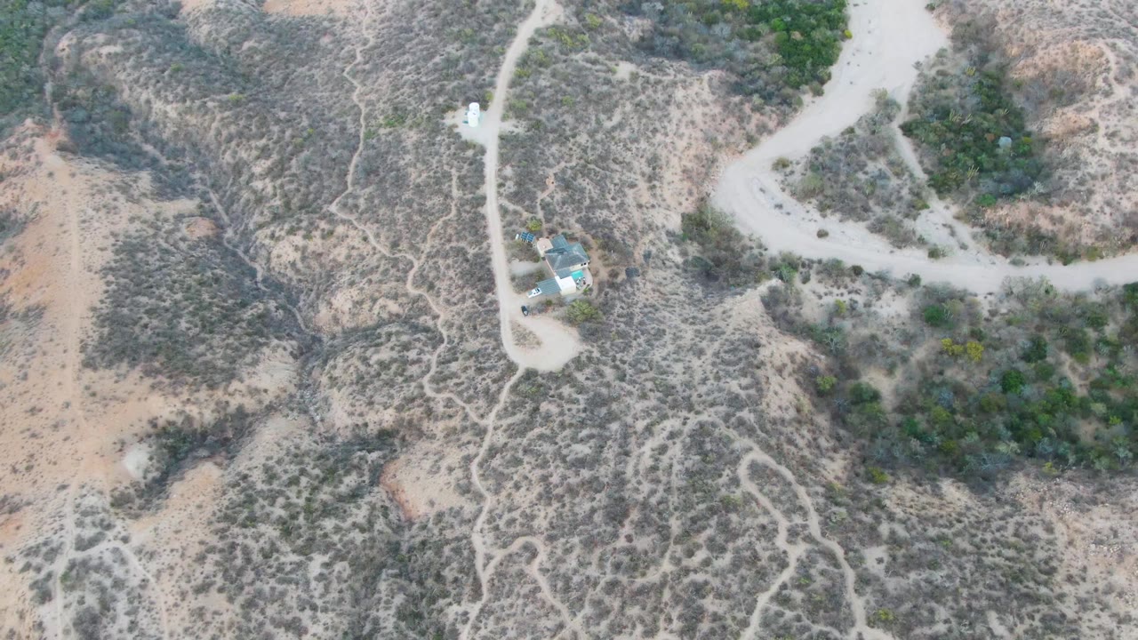 Overhead aerial frame of arid hillside terrain near Cabo Pulmo, Baja California Sur, with pale dirt roads, sparse scrub, small structures, and no visible people.