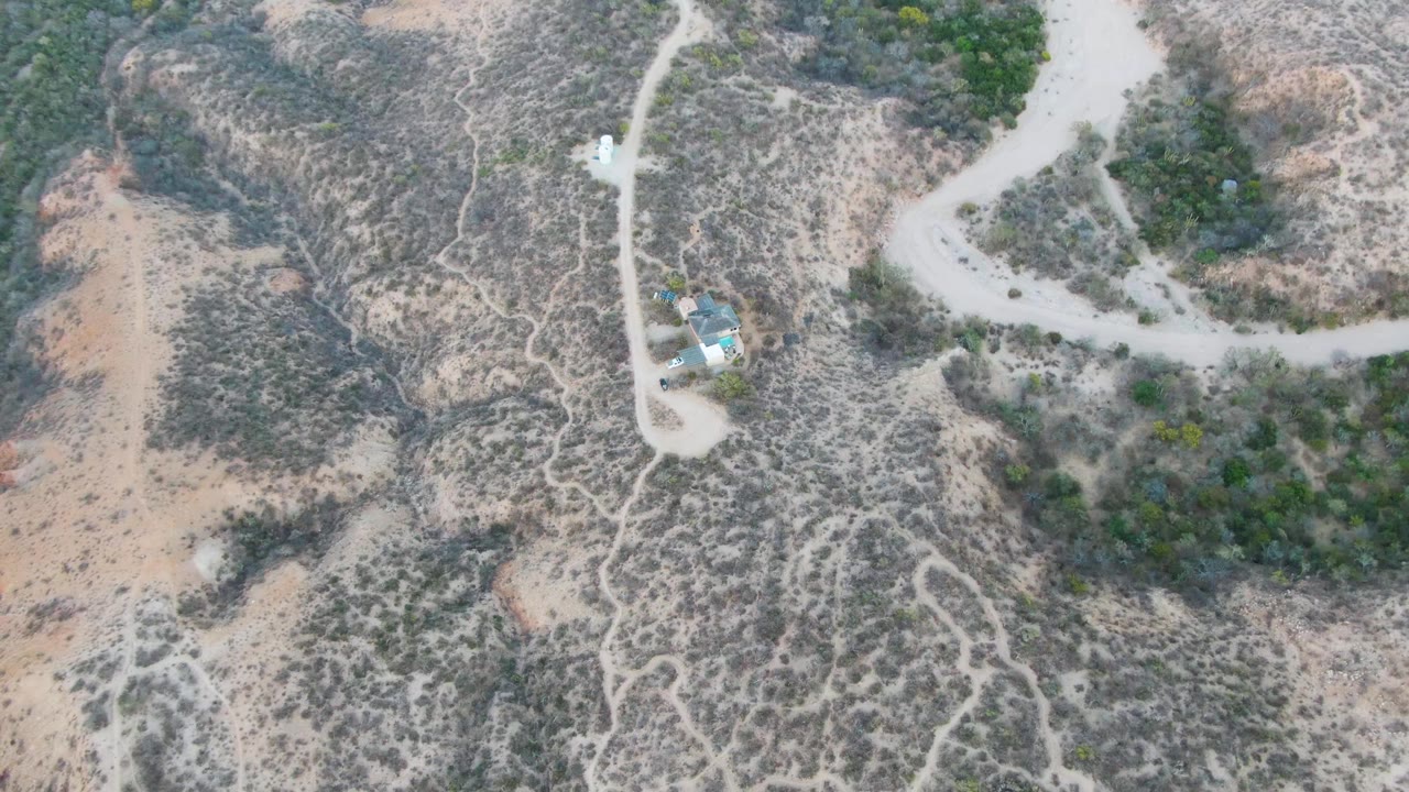 Aerial view over Cabo Pulmo dry hillside with a winding pale dirt road, scrub vegetation, rocky ground, and a small blue-roofed structure visible, with no visible people.
