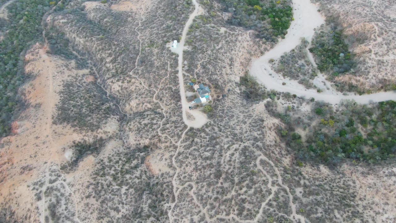 High aerial frame from Cabo Pulmo showing pale road bends across dry brown terrain, denser green scrub in one section, small structures, and no visible people.