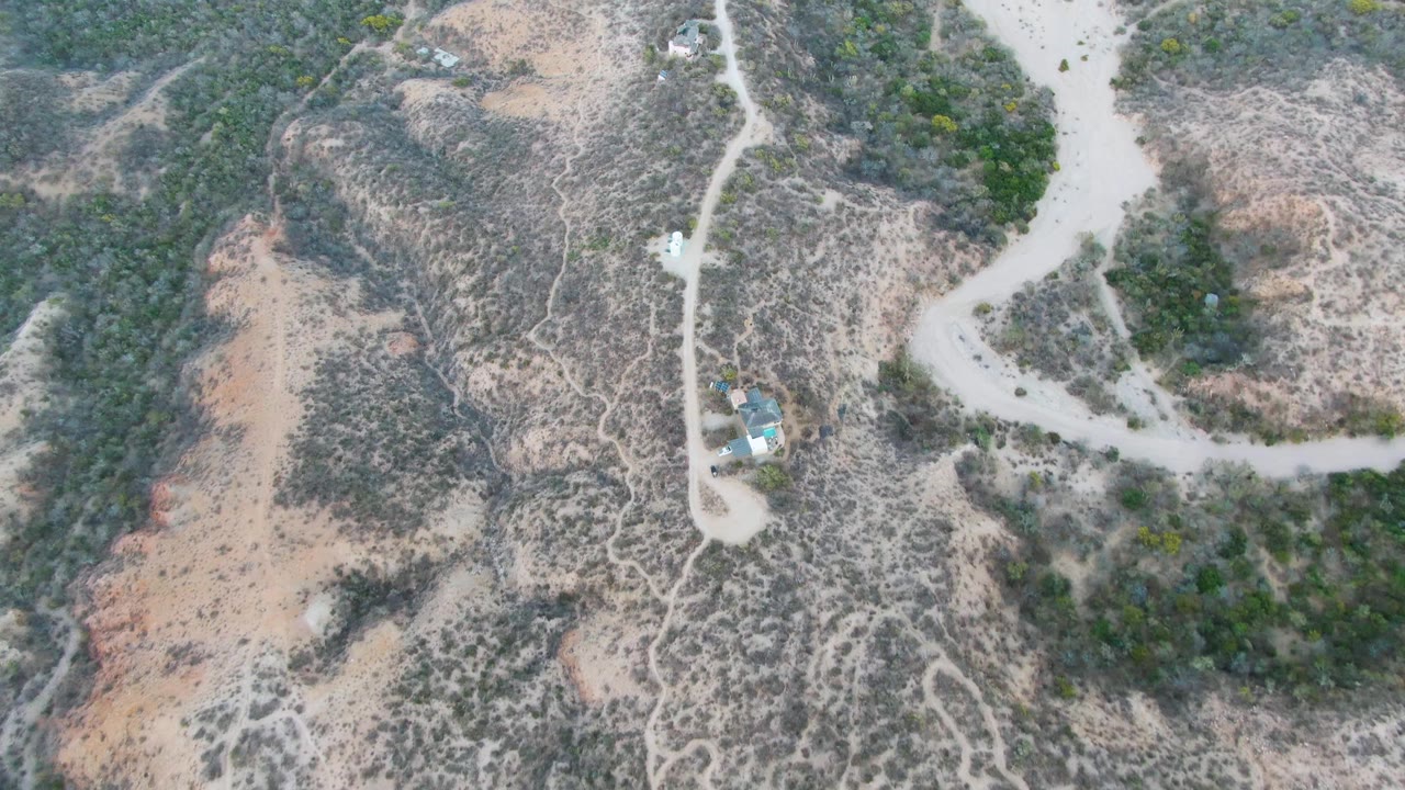 Aerial frame from Cabo Pulmo, Baja California Sur, with the vehicle slightly farther along the curved dirt road and dry brush covering the surrounding slopes, with no visible people.