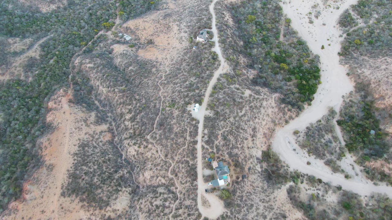 Aerial frame over Cabo Pulmo dry hills showing a pale vehicle on a winding dirt road, scrub-covered slopes, tan washes, a few small buildings, and no visible people.
