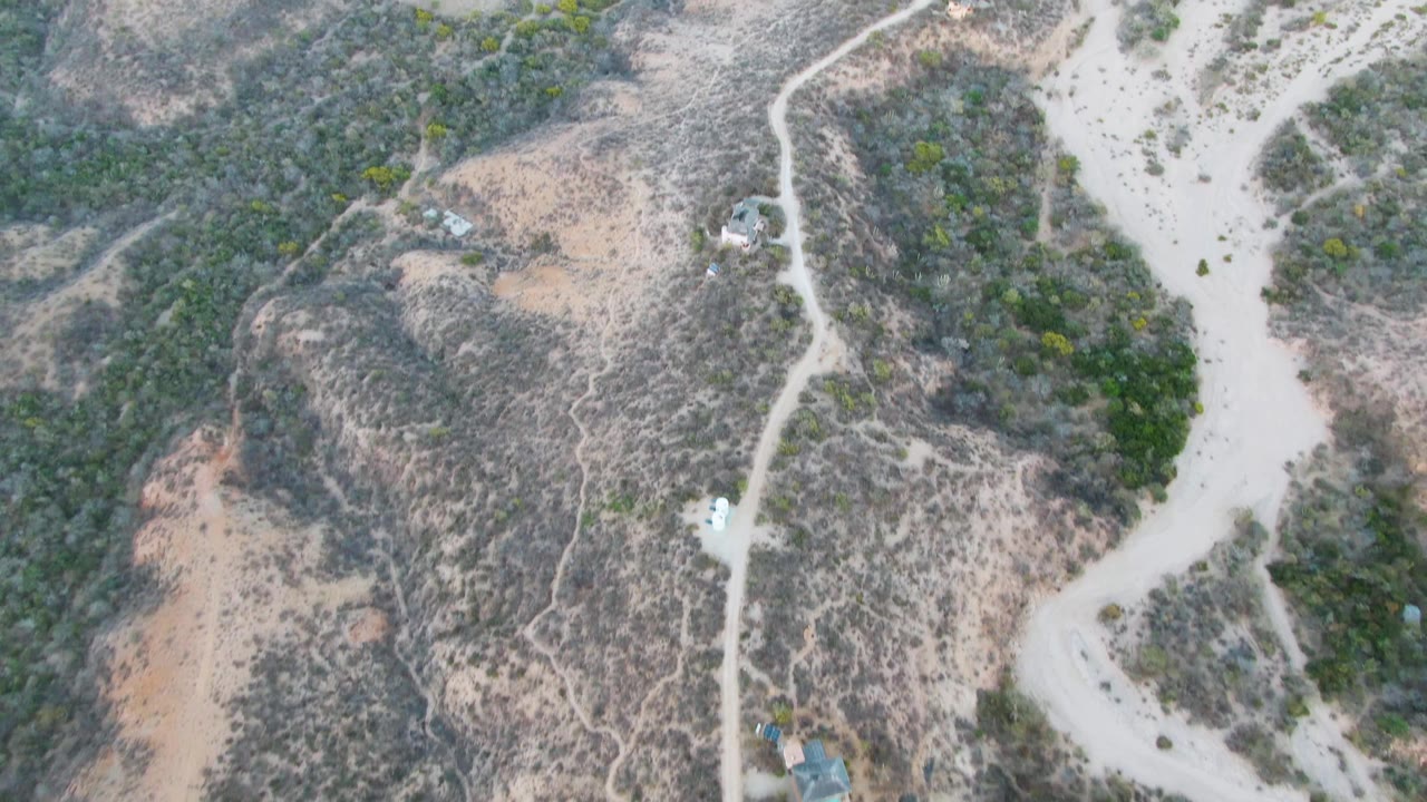 High aerial frame over Cabo Pulmo terrain where the dirt road curves between scrubby slopes and pale sandy washes, with the vehicle small in frame and no visible people.