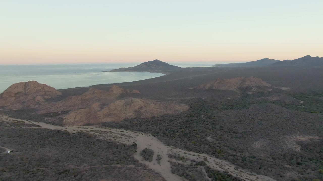 Aerial view in Cabo Pulmo with the dirt road cutting across scrubby foreground terrain, rounded rocky hills in midframe, coastal water behind them, and no visible people.