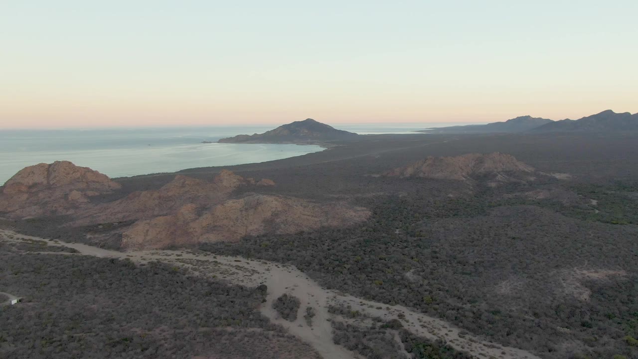 Cabo Pulmo aerial still showing a pale road through dark scrubland, tan rocky hills, a calm blue waterline, far mountains, pastel sunset color, and no visible people.