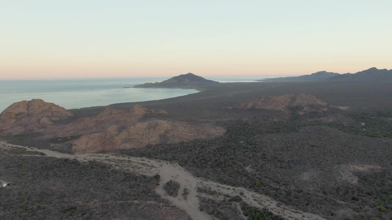 Wide aerial frame over Cabo Pulmo with dry foreground washes, the road bending through low vegetation, rocky hills near center, coastal water to the left, and no visible people.