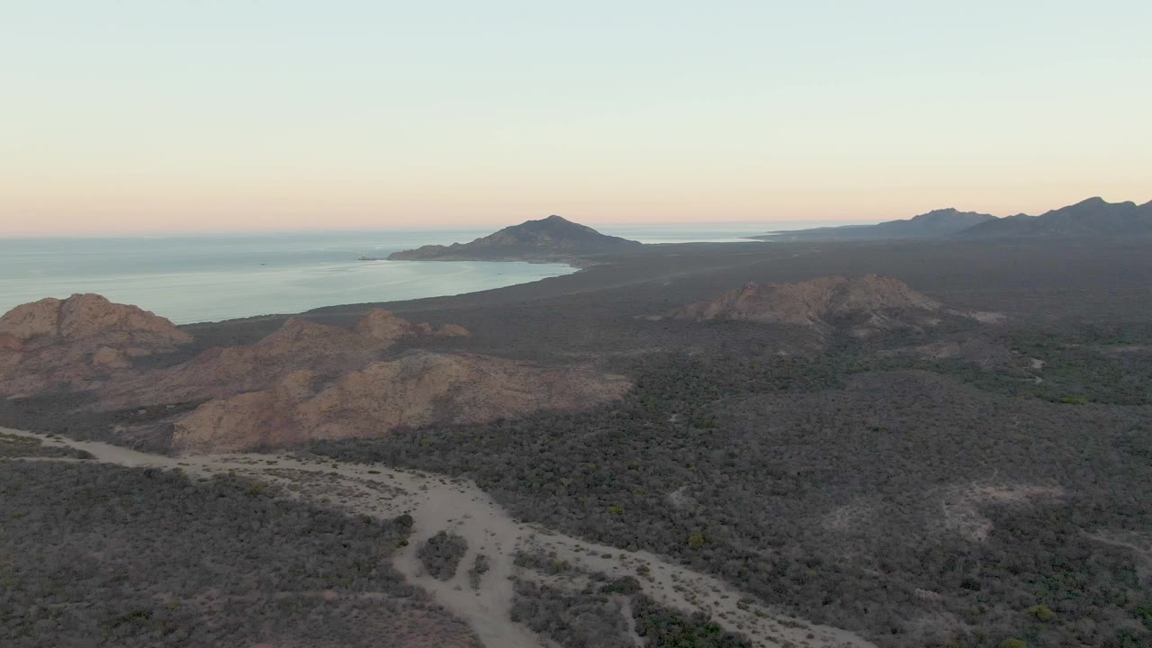 Aerial dusk view over Cabo Pulmo, Baja California Sur, with a pale dirt road curving through dry scrubland, rocky hills, distant ocean, and no visible people.