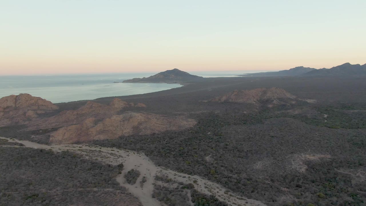 Aerial frame over Cabo Pulmo showing the dirt road lower in the frame, brown rocky hills across the middle distance, ocean to the left, and no visible people.