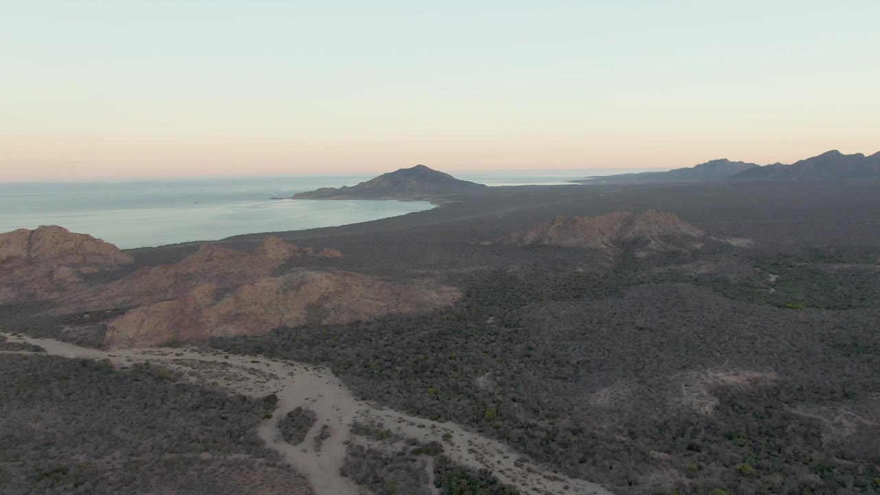 Aerial view in Cabo Pulmo with the road bend visible near the lower center, broad dry vegetation across the terrain, distant coastline at left, and no visible people.