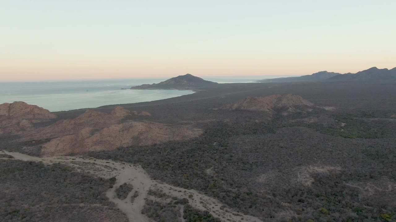 Aerial dusk view of a dirt track crossing dry hills toward a distant coastline.