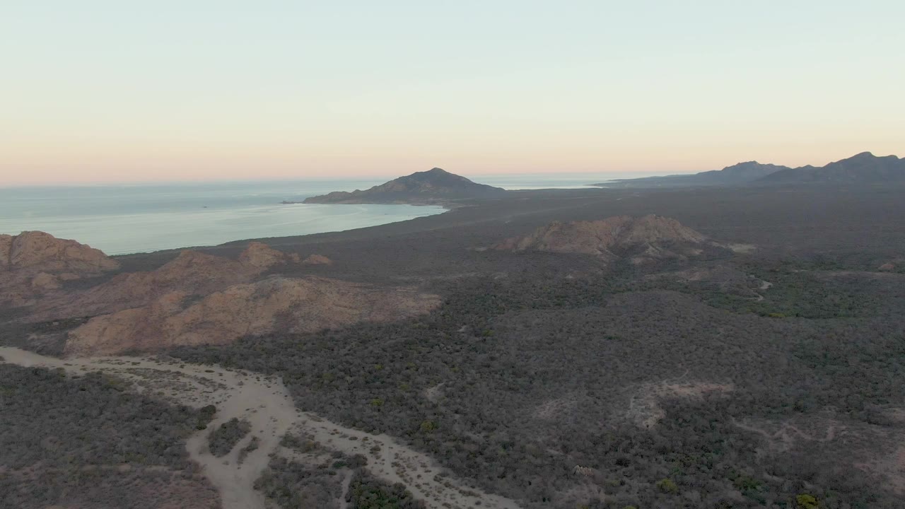 Aerial dusk view over Cabo Pulmo dry hills with a pale dirt road in the foreground, ocean to the left, distant mountains on the horizon, and no visible people.