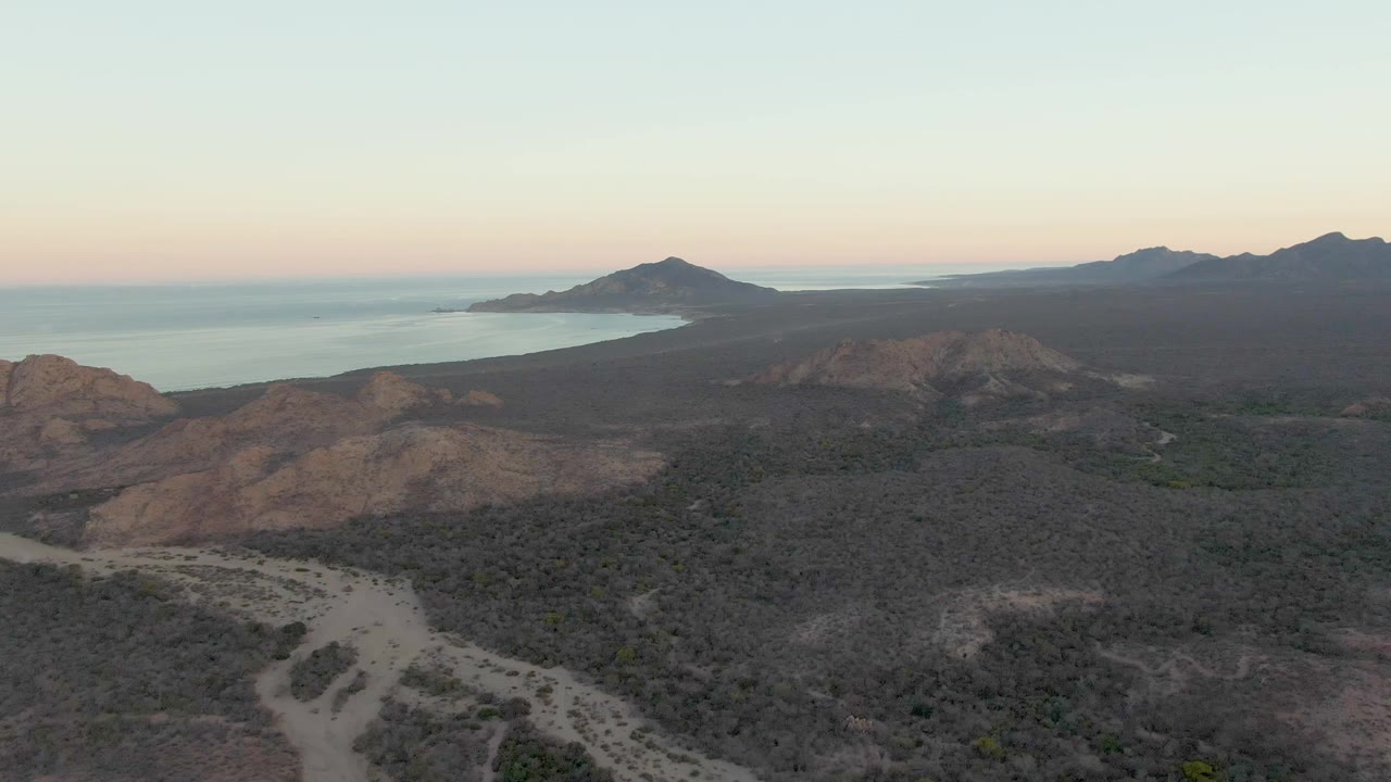 Wide aerial frame from Cabo Pulmo showing the dirt road curving across the lower terrain, scrubby hills in the middle distance, ocean at left, and no visible people.
