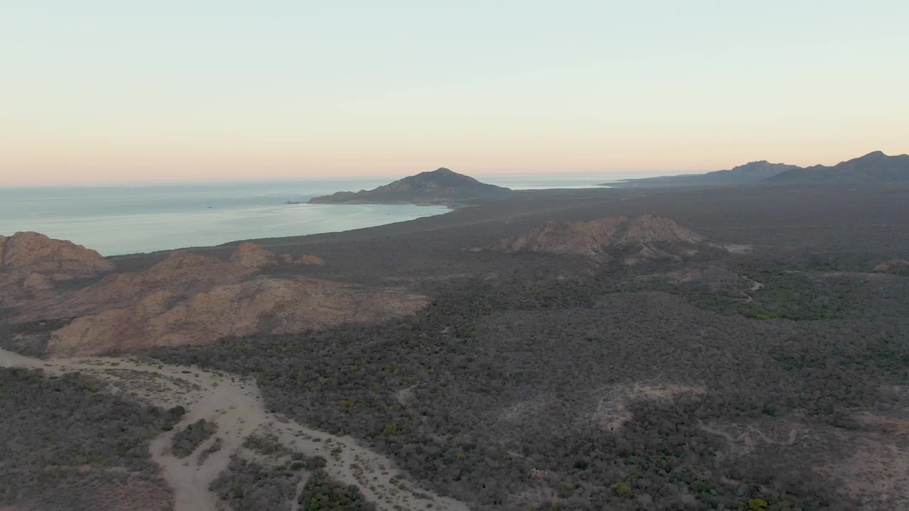Cabo Pulmo aerial view at dusk with a pale road cutting through dark scrub, rounded brown hills ahead, ocean visible on the left edge, and no visible people.