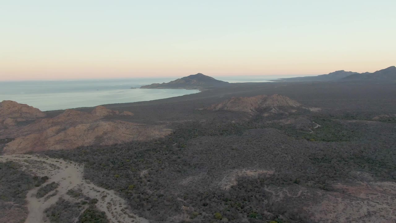 Aerial view near Cabo Pulmo showing dry ridges, dark scrubland, a pale ocean band, distant hills, and no visible people as the dusk light softens.