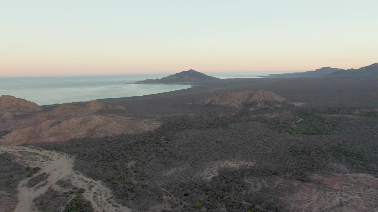 Wide aerial frame over Cabo Pulmo coastal terrain, with brown rocky hills below, darker scrub across the plain, ocean at left, distant mountains at right, and no visible people.