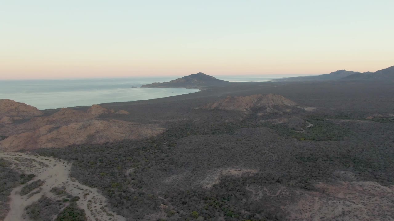 Dusk aerial frame in Cabo Pulmo with foreground rocky slopes, dense-looking low scrub, a blue-gray ocean strip, dark distant mountains, and no visible people.