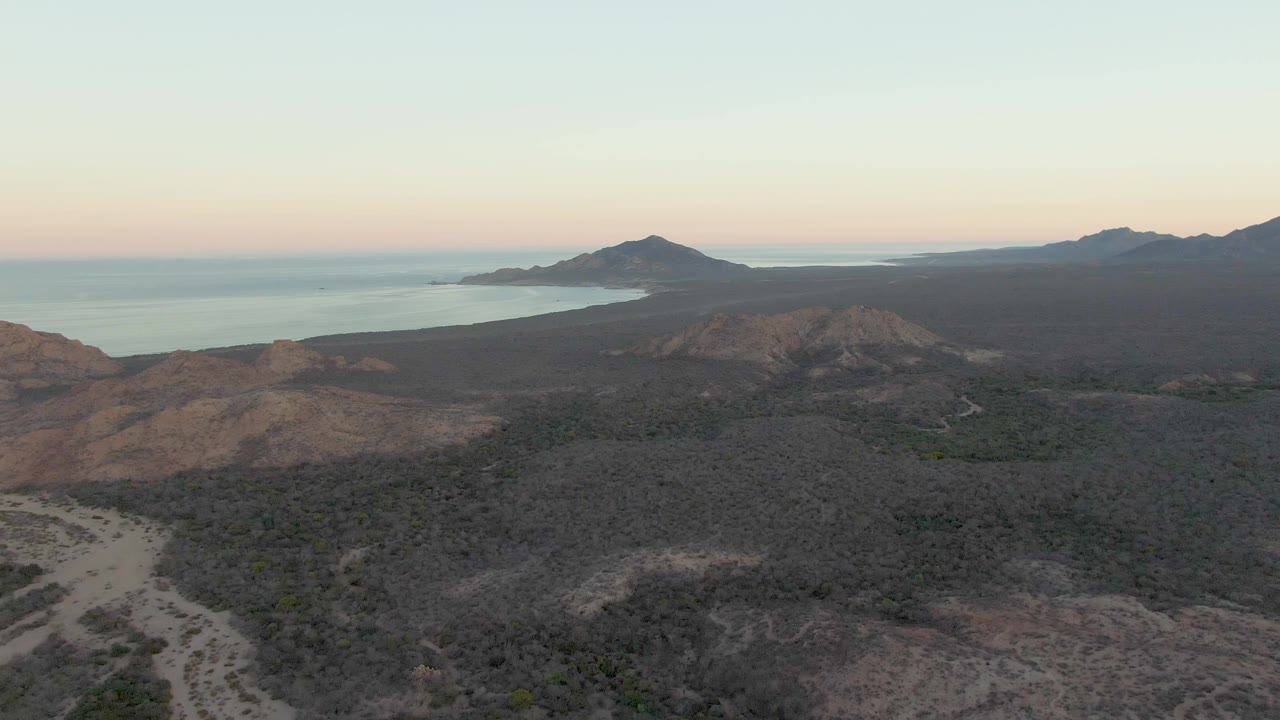 Aerial frame over Cabo Pulmo desert terrain shows a pale winding road crossing low scrub hills toward a calm ocean, distant mountains, soft dusk sky, and no visible people.