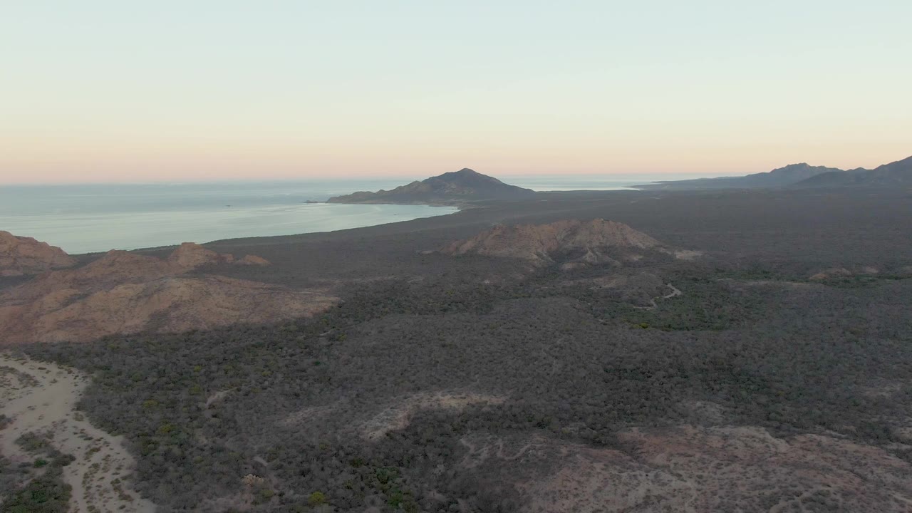 At 0:02, the aerial composition over Cabo Pulmo shows dry ridges and sparse vegetation in front, a winding road with a small vehicle, ocean to the left, mountains on the horizon, and no visible people.