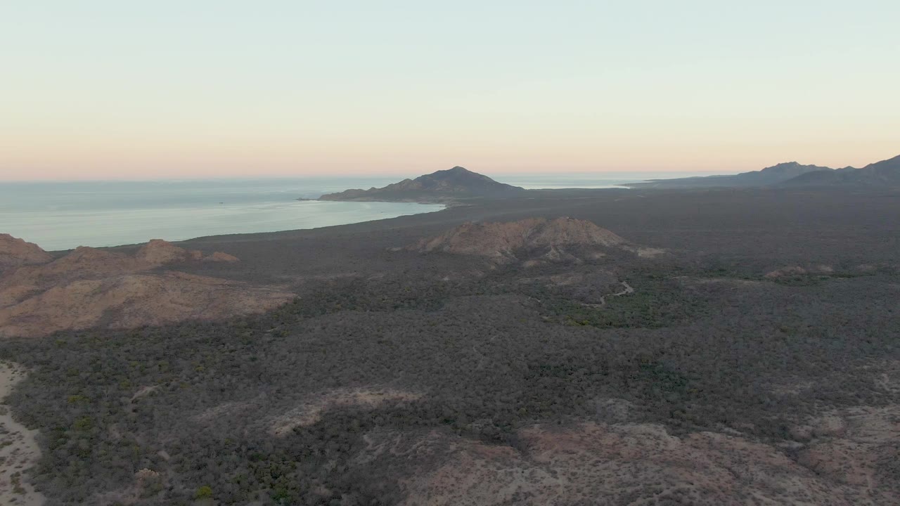 Aerial view over dark volcanic terrain in Cabo Pulmo, Baja California Sur, with a faint pale road below, ocean behind the hills, distant mountains, and no visible people.