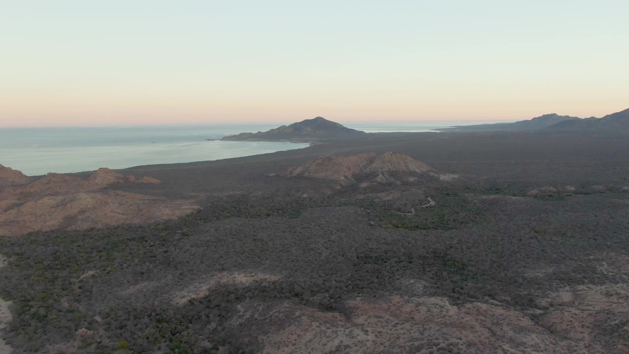 Aerial frame from Cabo Pulmo showing dry rocky hills in the foreground, the road receding near the lower center, calm ocean in the background, mountain silhouettes, and no visible people.