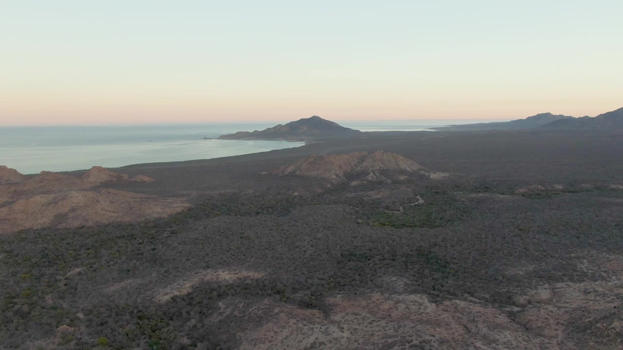 Wide aerial view in Cabo Pulmo with dark scrub-covered volcanic ground across the foreground, a faint road thread lower in frame, ocean water beyond, distant ridges, and no visible people.