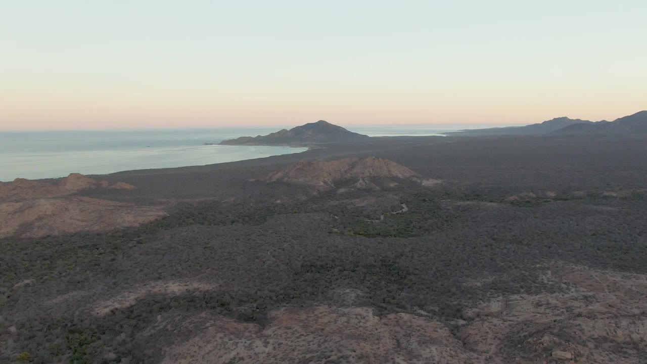 Aerial frame over Cabo Pulmo showing dark rocky lava field in the foreground, low hills, calm ocean, a distant island, and no visible people.