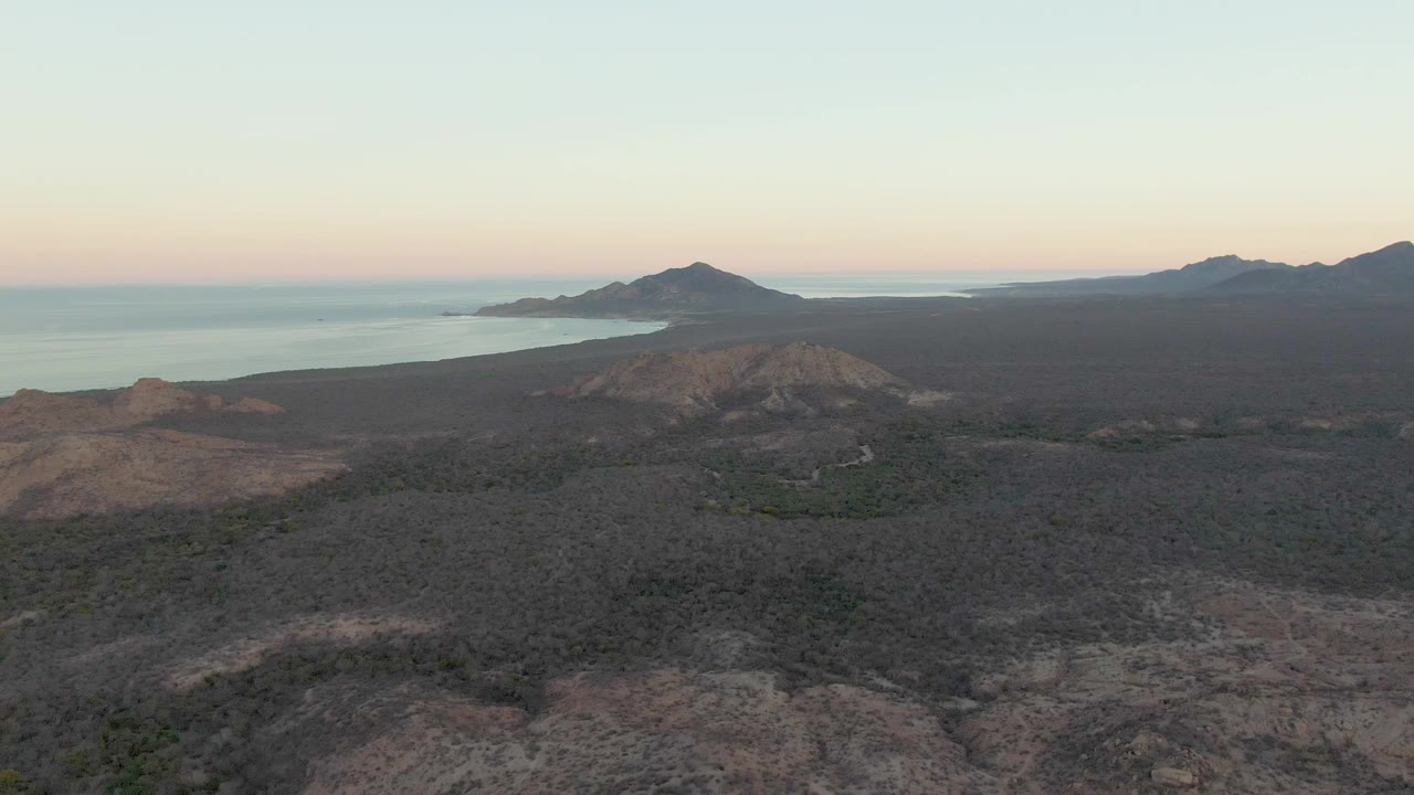 Second-frame aerial over Cabo Pulmo with a broad dark lava field below, scrub patches, a rounded hill, distant ocean, island silhouettes, and no visible people.