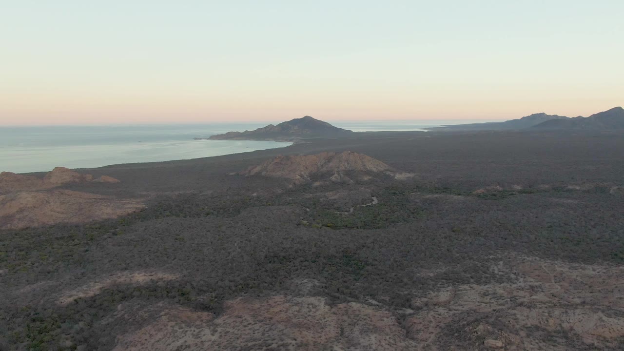 Mid-clip aerial frame in Cabo Pulmo showing dark scrubland, brown volcanic rises, calm water beyond, a distant island ridge, and no visible people.